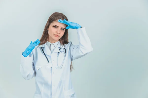 Atrakcyjna stażystka w białym płaszczu medycznym i stetoskopie na szyi, stojąca, trzymająca rękę na czole, jakby kogoś szukała, zawężająca oczy. — Zdjęcie stockowe