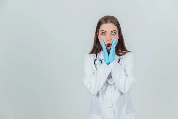 Vrouwelijke stagiaire in medisch uniform, houdt handen op de wangen, kijkt naar de camera doodsbang en bang, mond en ogen wijd open. — Stockfoto