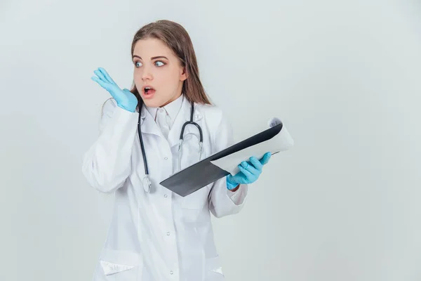 Young female intern standing in medical uniform, holding clipboard. Shocked by what she found out. — ストック写真