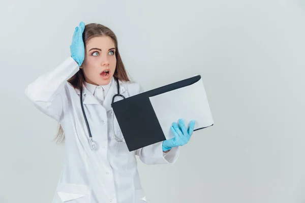 Young female intern standing in medical uniform, holding clipboard. Shocked by what she found out, keeping hand on the head. — ストック写真