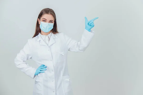 Portret młodej lekarki w masce medycznej, sterylnych rękawiczkach i patrzącej w kamerę, patrzącej palcem w przestrzeń do kopiowania. — Zdjęcie stockowe