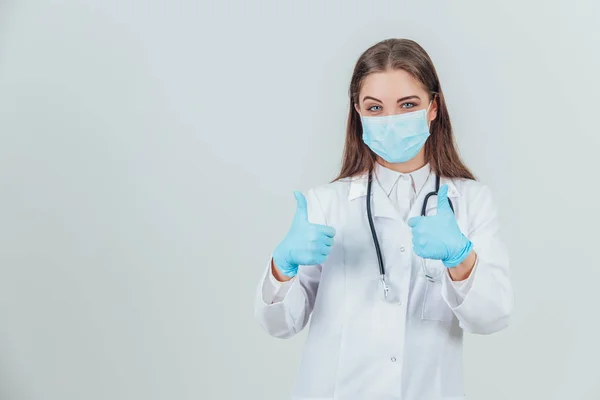 Portrét mladého doktora v lékařské masce, na sterilních rukavicích a se stetoskopem a při pohledu do kamery, ukazující palce nahoru. — Stock fotografie