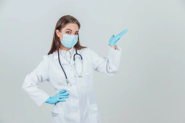 Portret młodego lekarza w masce medycznej, na sterylnych rękawiczkach i stetoskopie, patrząc w kamerę, trzymając ręce akimbo, wskazując na przestrzeń na boku. — Zdjęcie stockowe