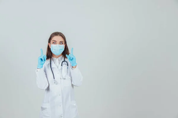 Portrét mladého doktora v lékařské masce, na sterilních rukavicích a se stetoskopem, ukazuje prsty nahoru. — Stock fotografie