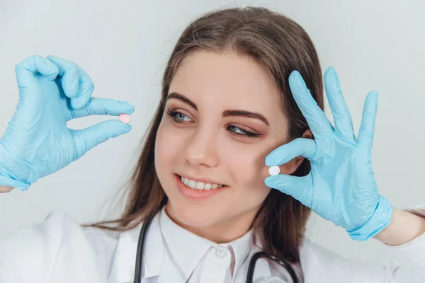 Krásná, usmívající se mladá doktorka s dvěma malými bílými pilulkami v prstech u obličeje, dívá se na jednu z nich, usmívá se. — Stock fotografie