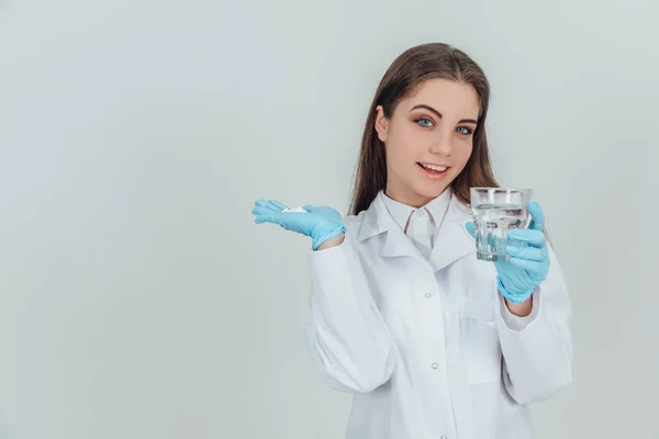 Ładna kobieta w białym płaszczu trzyma stertę tabletek w jednej ręce, a szklankę wody w drugiej. Gotowy do stosowania leku. — Zdjęcie stockowe