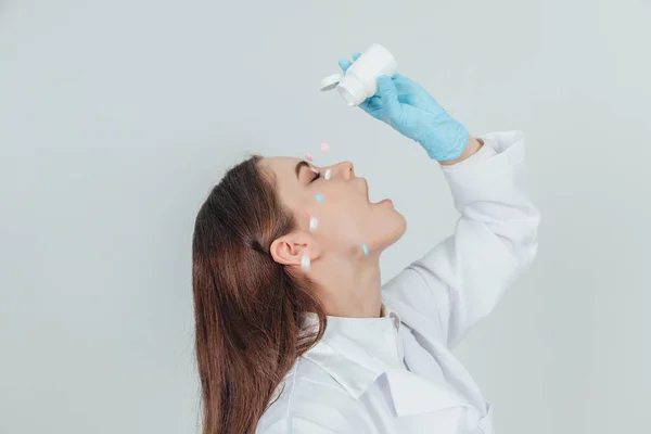 Vrij lachende stagiaire meid die pillen uit de witte fles op haar gezicht giet. Ogen dicht, mond open. De pillen vallen overal.. — Stockfoto