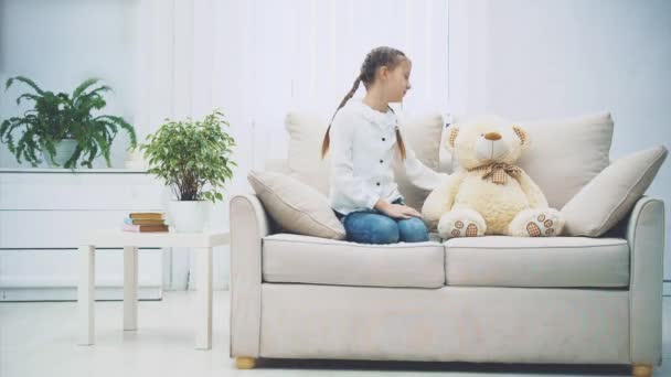 Charmerande unge sitter i soffan med benen hoprullade och pratar med nån vid sidan om. Vit teddybjörn är bredvid henne.. — Stockvideo