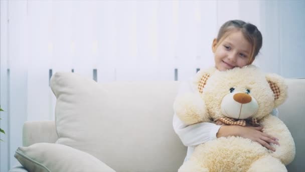 Ευτυχισμένο κοριτσάκι που κάθεται στον καναπέ, αγκαλιάζοντας ένα μεγάλο αρκουδάκι.. — Αρχείο Βίντεο