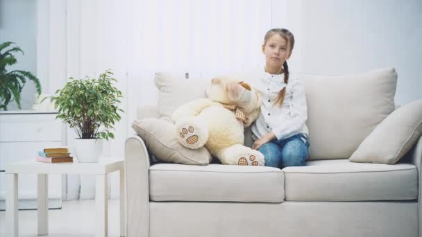 小女孩坐在沙发上，看起来心烦意乱，无聊极了，然后抱着一只大泰迪熊笑了。 永远的朋友. — 图库视频影像