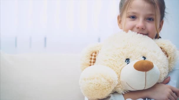 Niña feliz sentada en el sofá, abrazando a un gran osito de peluche . — Vídeos de Stock