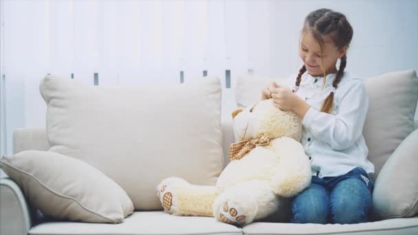 Menina bonita sentada no sofá, brincando com seu ursinho de pelúcia, tocando seu nariz e ouvidos . — Vídeo de Stock