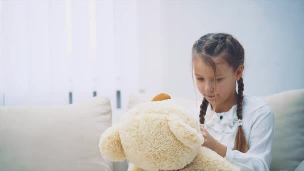 Hübsches Mädchen sitzt auf dem Sofa, spielt mit ihrem Teddybär und justiert seine Fliege neu. — Stockvideo