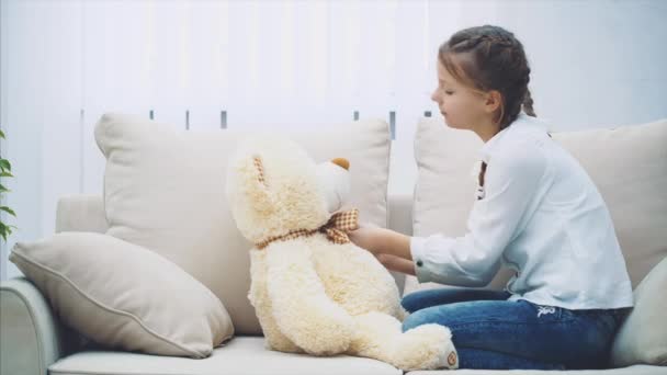 Mooi meisje zitten op de bank, spelen met haar teddybeer, bijstellen van haar strikje. — Stockvideo