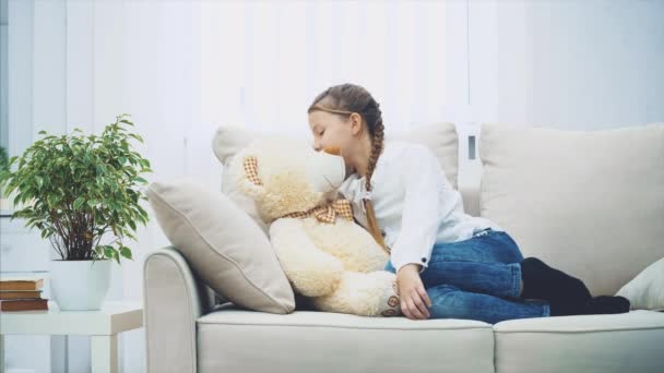 Roztomilá holčička sedí na gauči, povídá si se svým medvídkem a šeptá mu do ucha nějaké tajemství. — Stock video