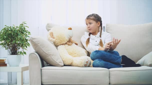 Χαριτωμένο κοριτσάκι που κάθεται στον καναπέ, μιλάει με το αρκουδάκι της, λυγίζοντας τα δάχτυλά της απελπισμένα.. — Αρχείο Βίντεο