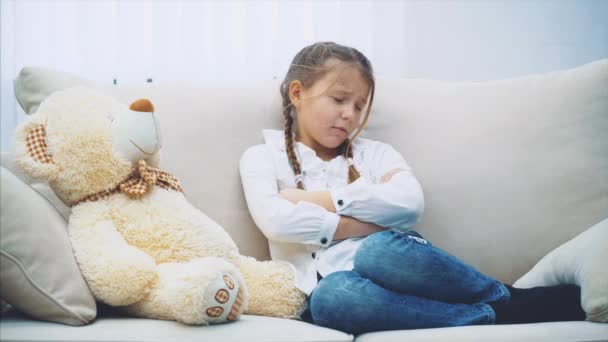 Linda niñita sentada en el sofá, hablando con su osito de peluche, explicando desesperadamente, sabiendo que no la entiende pero abrazando a este gran osito de peluche . — Vídeos de Stock