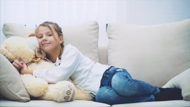 Sevimli küçük kız kanepede uzanıyor, büyük oyuncak ayısına sarılıyor.. — Stok video