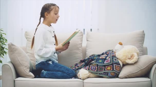 Linda niña sentada con las piernas acurrucadas, leyendo un cuento de hadas para su peluche, que yace debajo de la manta . — Vídeo de stock