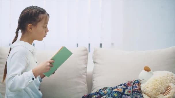 Schattig klein meisje zitten met haar benen opgerold, het lezen van sprookje voor haar teddy, dat ligt onder de deken. — Stockvideo