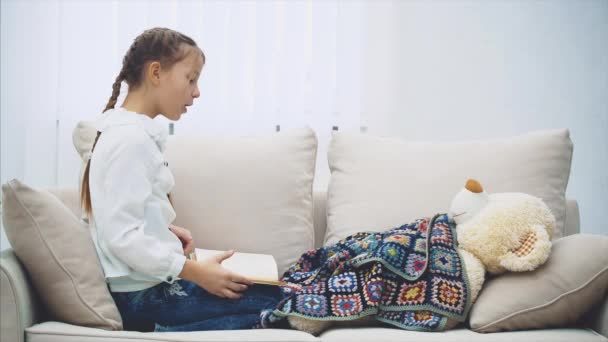 Gadis kecil yang lucu duduk dengan kaki meringkuk, membaca dongeng untuk boneka teddy-nya, menggerakkan tangannya emosional, menunjukkan plot buku . — Stok Video