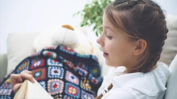 Gadis kecil yang lucu duduk dengan kaki meringkuk, membaca dongeng untuk boneka teddy-nya, menggerakkan tangannya emosional, menunjukkan plot buku . — Stok Video