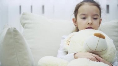 Küçük sevimli kız kanepede oturuyor ve büyük peluş arkadaşına sarılıyor, iç çekiyor. Üzgün ve endişeli görünüyor..