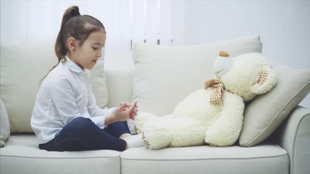 Cute little girl sitting on the sofa, talking with her teddy, bending her fingers desparately, shaking teddy. — ストック動画