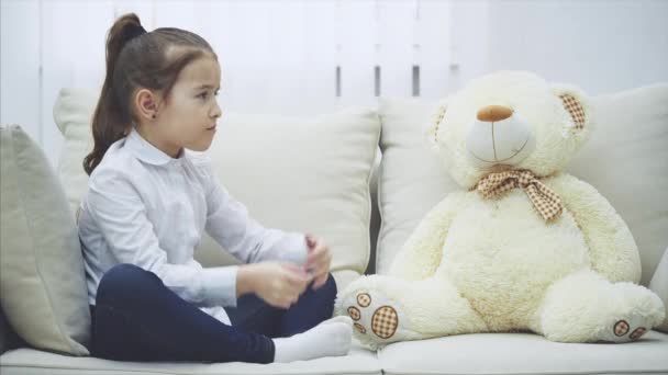 Симпатична маленька дівчинка сидить на дивані, розмовляє зі своєю плюшевою, згинаючи пальці, дивлячись розчаровано . — стокове відео
