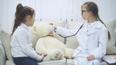 Oyuncak ayıyla oynayan iki tatlı çocuk. Doktoru oynayan kız peluş ayıyı steteskopla muayene ediyor, başka bir kızla konuşuyor, ayıya hap veriyor..