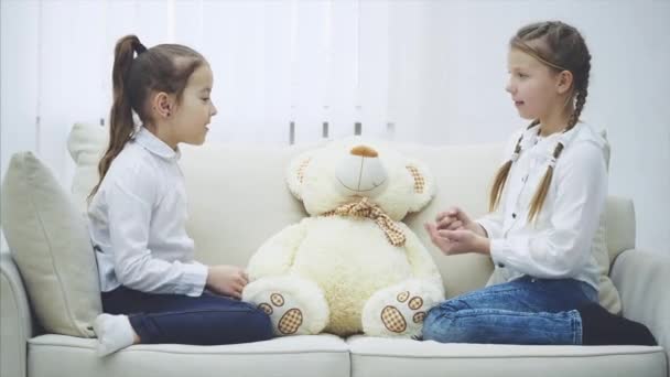 Zwei hübsche Mädchen, die auf dem Sofa sitzen und Steinpapier-Scheren spielen, um zu entscheiden, wer mit dem weißen weichen Bären spielen wird. — Stockvideo