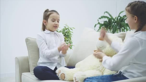 Zwei hübsche Mädchen, die auf dem Sofa sitzen und Steinpapier-Scheren spielen, um zu entscheiden, wer mit dem weißen weichen Bären spielen wird. — Stockvideo