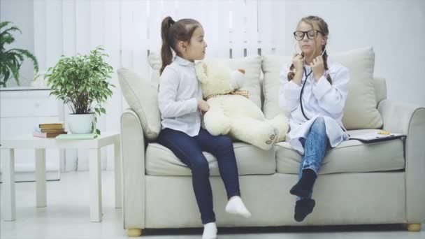 Dos dulces niños jugando con un osito de peluche. La chica que interpreta a un médico está examinando al oso de peluche con estetoscopio. . — Vídeo de stock