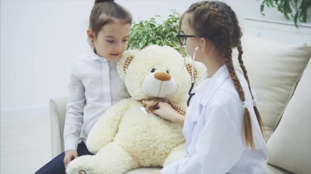 Два милых ребенка играют с плюшевым мишкой. Девушка, играющая доктора, осматривает плюшевого медведя стетоскопом, разговаривает с другой девушкой, пишет диагноз . — стоковое видео