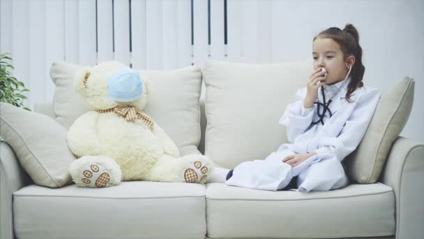 Little girl in medical coat is talking in the stethoscope. White teddy-bear in medical mask is sitting near her. — Stock Video