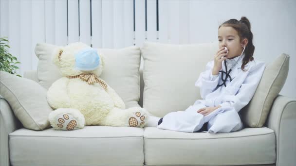 Az orvosi köpenyes kislány a sztetoszkópban beszél. Fehér maci orvosi maszkban ül mellette..