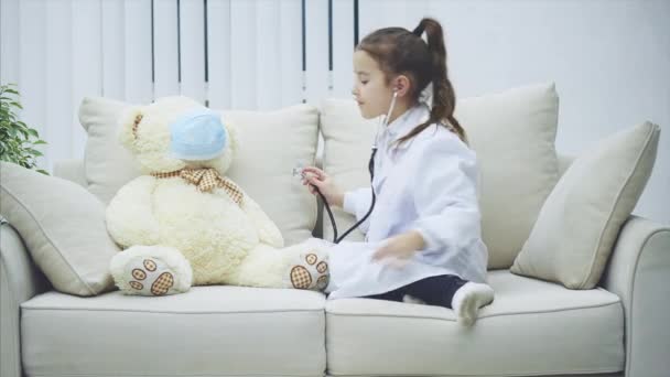 Little girl in medical coat is examining teddy-bear with the stethoscope, then talking with it. — Stock Video