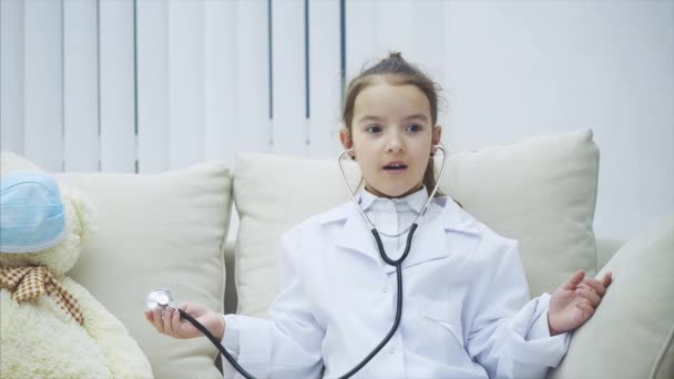 La niña con abrigo médico se está examinando a sí misma, luego el osito de peluche con el estetoscopio. Expresión facial sorprendida . — Vídeos de Stock