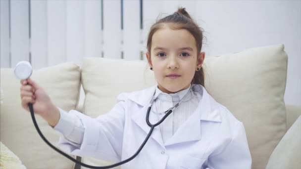 Menina engraçada está sentada no sofá com estetoscópio nos ouvidos examinando algo no ar . — Vídeo de Stock