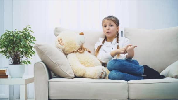 Menina bonito sentado no sofá, conversando com seu ursinho de pelúcia, explicando desparately, sabendo que não entende ela, mas abraçando este grande ursinho de pelúcia . — Vídeo de Stock