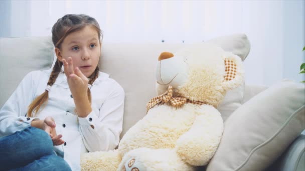 かわいい女の子はソファの上に座ってテディと話して指を曲げ. — ストック動画