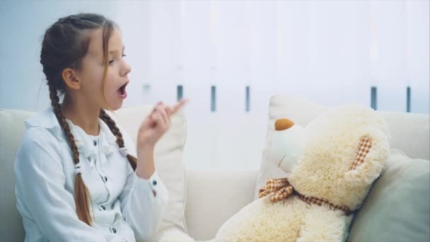 Gadis kecil yang lucu duduk di sofa, berbicara dengan boneka teddy-nya, memaki-maki itu, menggoyangkan jarinya, melipat tangannya, tampak kecewa bahwa itu tidak mengerti dirinya . — Stok Video