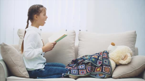 Niedliches kleines Mädchen sitzt mit zusammengerollten Beinen und liest Märchen für ihren Teddy, der unter der Decke liegt. — Stockvideo