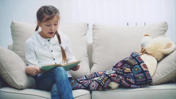 Linda niña sentada en el sofá, leyendo cuentos de hadas para su peluche, que está debajo de la manta . — Vídeo de stock