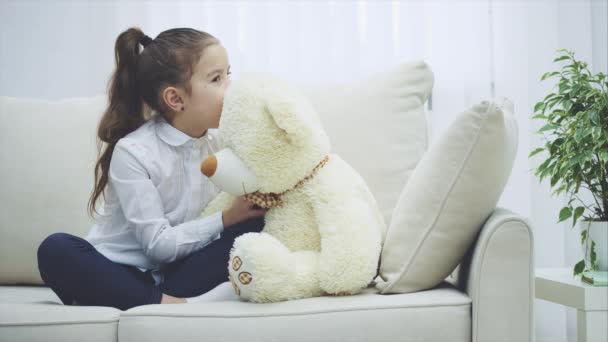 Щаслива маленька дівчинка сидить на дивані, обіймаючи великого плюшевого ведмедя . — стокове відео