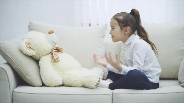 Cute little girl siedzi na kanapie, rozmawiając z jej misia, zginając jej palce desperacko. — Wideo stockowe