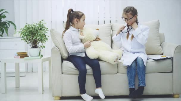 Dos dulces niños jugando con un osito de peluche. Una chica es doctora con estetoscopio. Ella está lista para escuchar al oso. . — Vídeo de stock