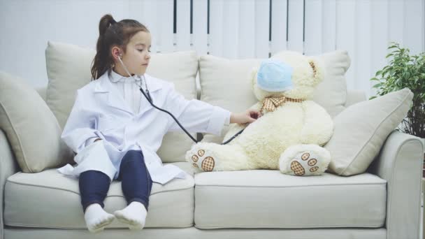 La niña está jugando con su peluche. Ella es doctora. El oso es un paciente. Chica está examinando su peluche con estetoscopio . — Vídeo de stock