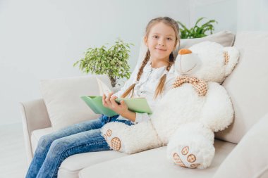 Kanepede oturan sevimli küçük kız yanında oyuncak ayıyla peri masalı okuyor, gülümsüyor..