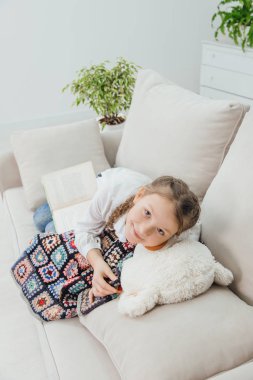 Güzel örgüleri olan gülümseyen küçük bir kız kanepesinde, oyuncak ayısının yanında, battaniyenin altında yatıyor..